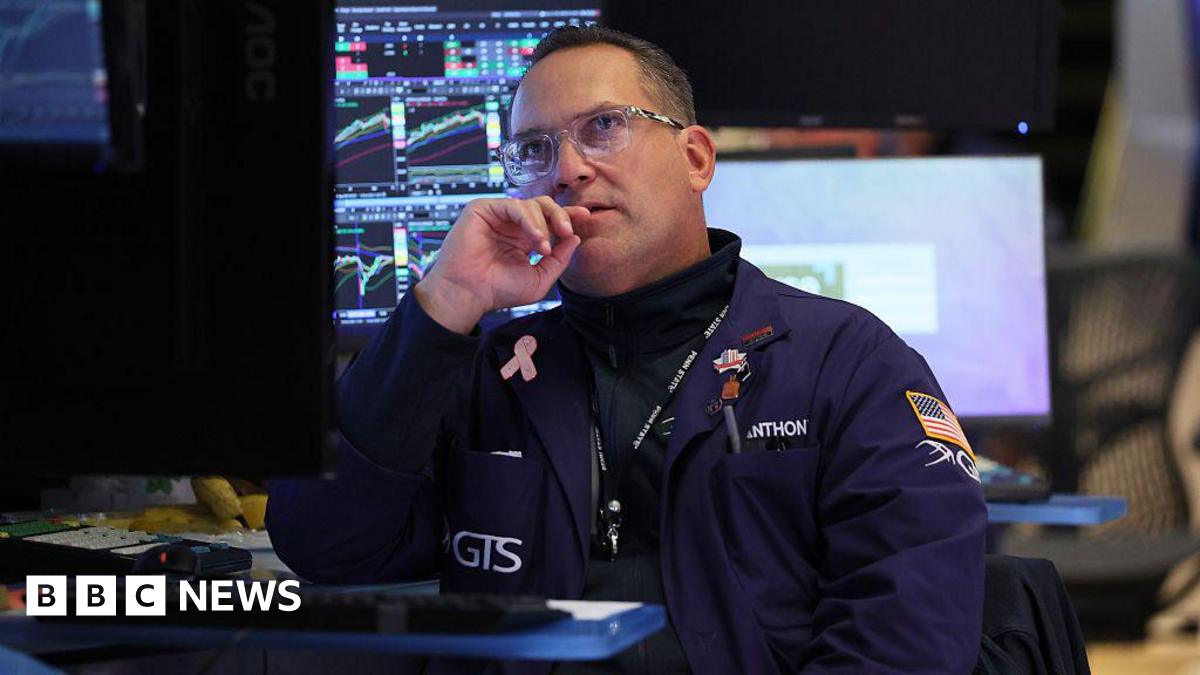 A trader in a dark blue coat with an American flag on his sleeve sits near computers at the New York Stock Exchange in October 2025, his eyes lifted to the screens above him.