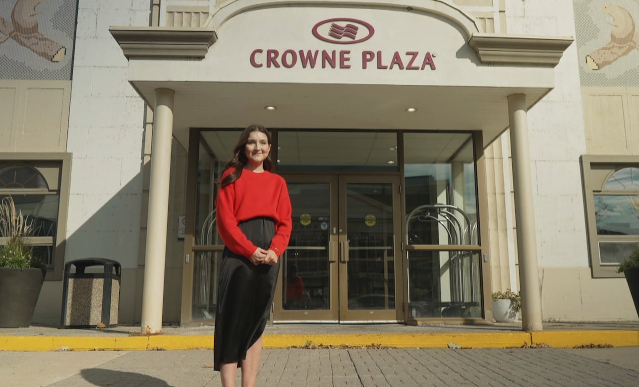 A woman wearing a red sweater and black skirt stands outside a hotel with the words "Crowne Plaza" on the front.