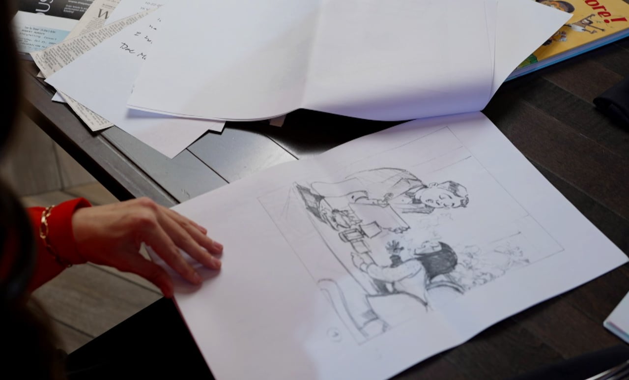 A woman's hand flipping through a booklet of pages. The opened page is a pencil drawing of a young girl speaking to a man over a dinner table.