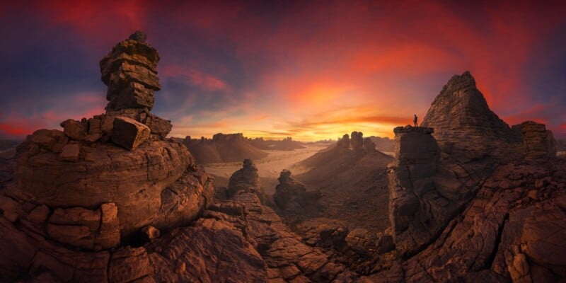 A dramatic rocky landscape at sunset, with jagged cliffs and formations under a vibrant orange and purple sky. A lone person stands on a rock ledge, admiring the vast, rugged terrain.