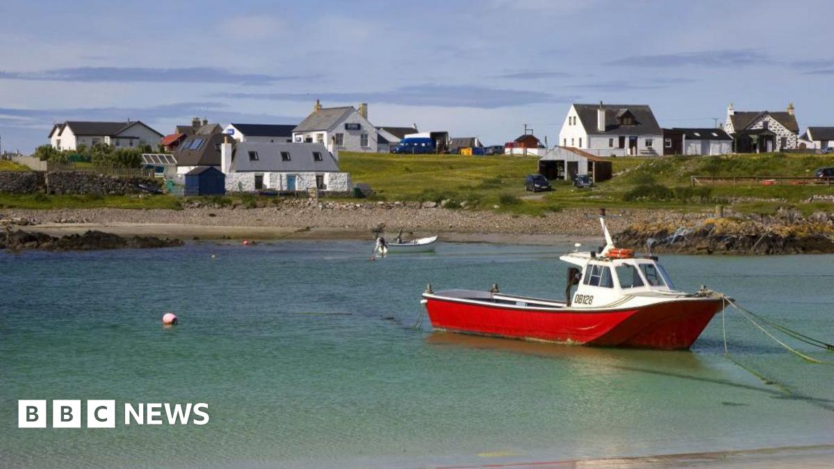 A small fishing boat with a red hull is tied up in a shallow expanse of turquoise water. Land in the background is dotted with white houses.