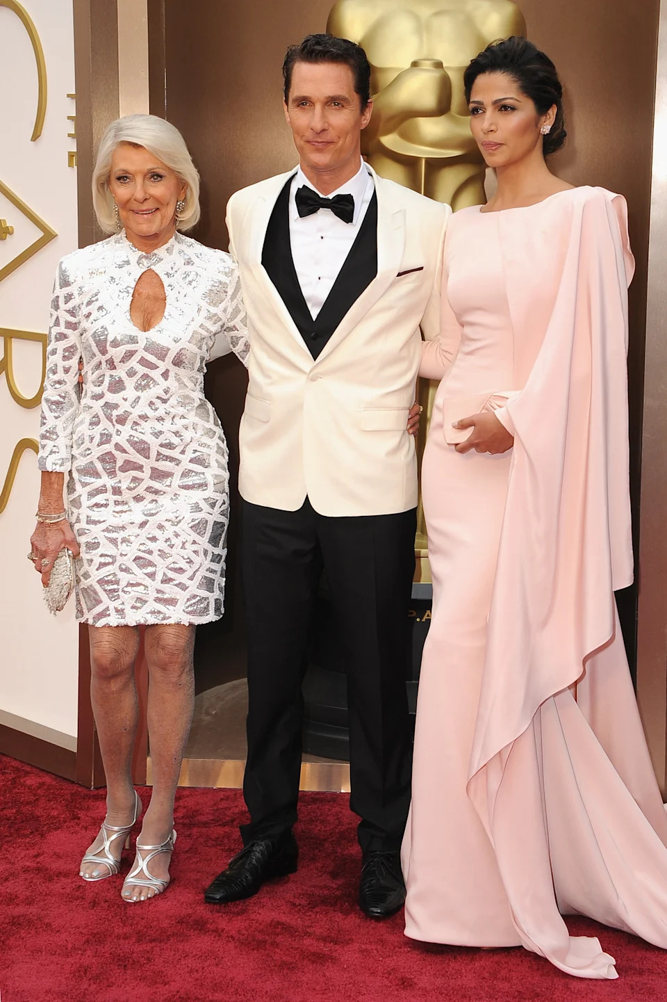 Three people pose elegantly on a red carpet. The man wears a white tuxedo, flanked by women in chic gowns with intricate designs