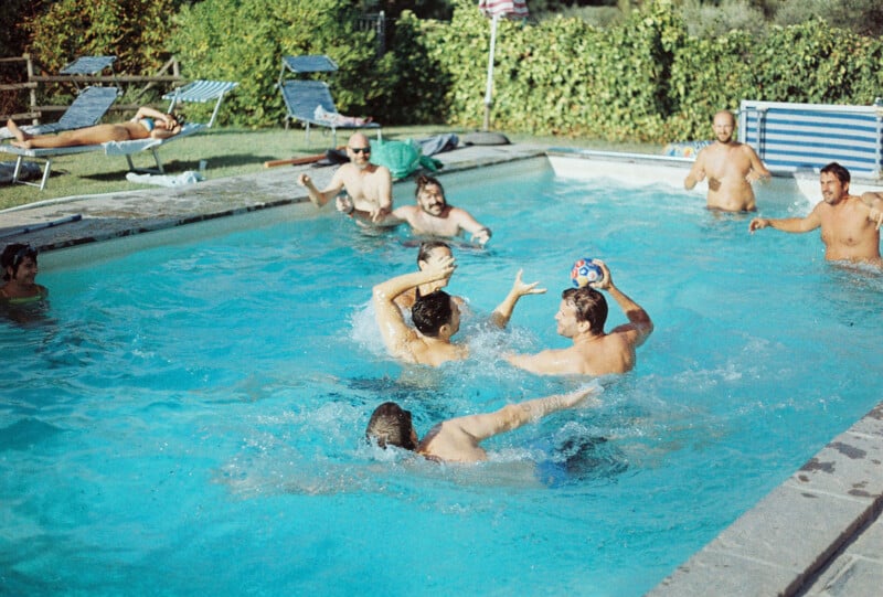A group of people play and splash with a ball in an outdoor swimming pool on a sunny day; others relax on lounge chairs nearby.