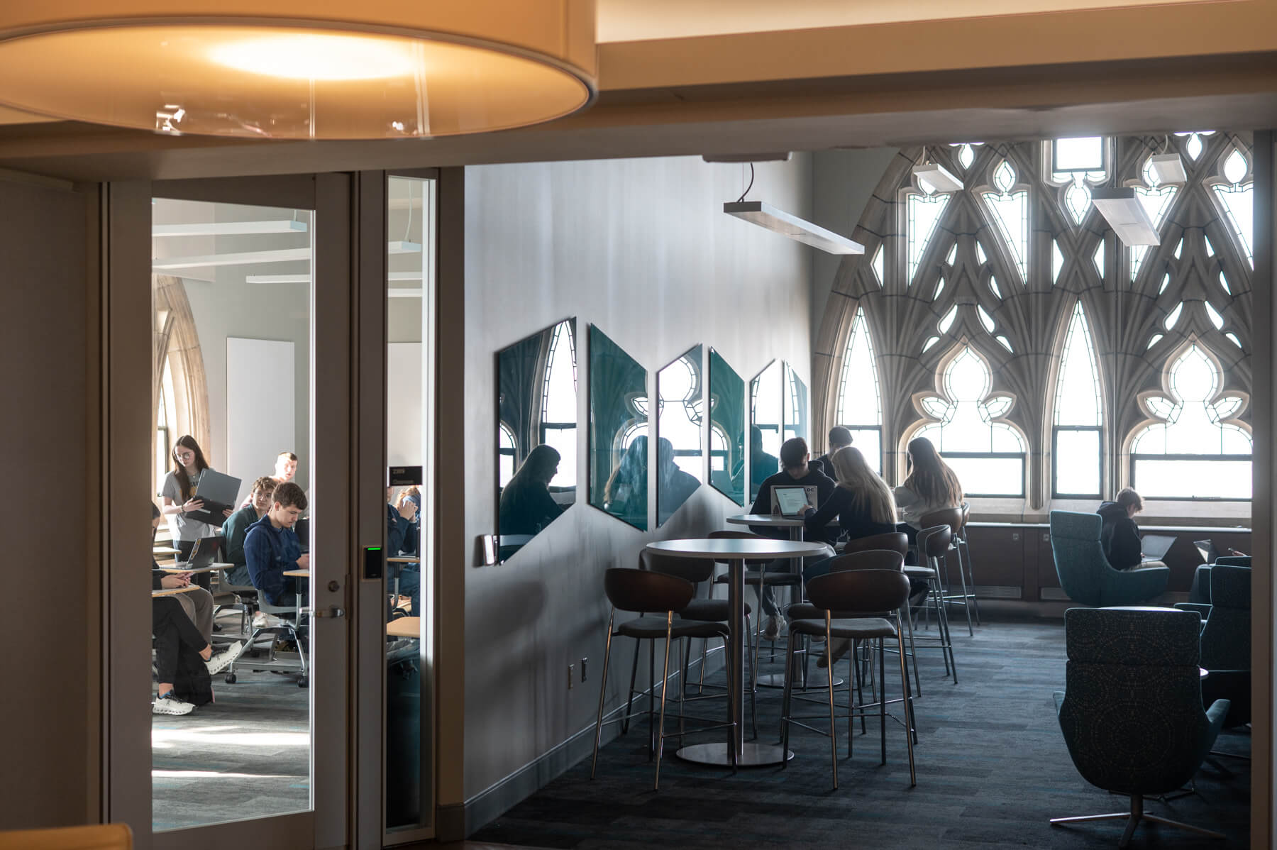 People work in a modern office space with large arched windows, seated at desks and high tables, with visible reflections on hexagonal mirrors on the wall.