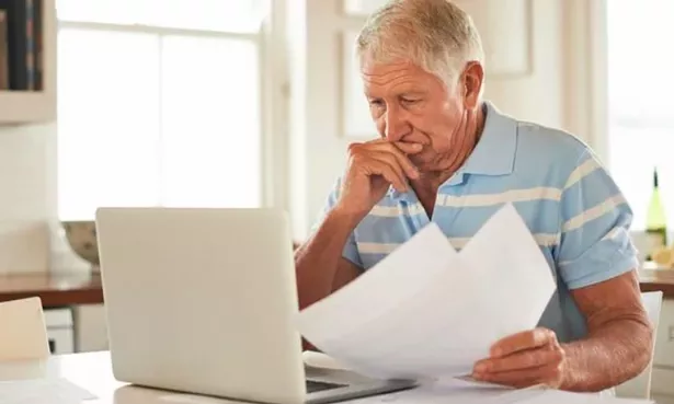 An older man is sitting looking over paperwork with a pensive expression on his face