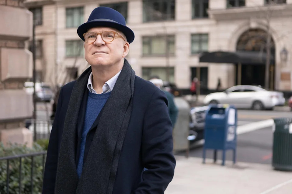 Steve Martin stands on a sidewalk, wearing a fedora, glasses, a dark coat, and scarf. A building and parked cars are in the background