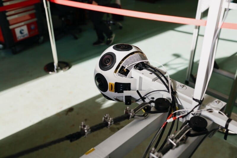 A spherical device with multiple cameras mounted on a metal frame in an indoor industrial setting, with cables attached and various equipment and barriers in the background.