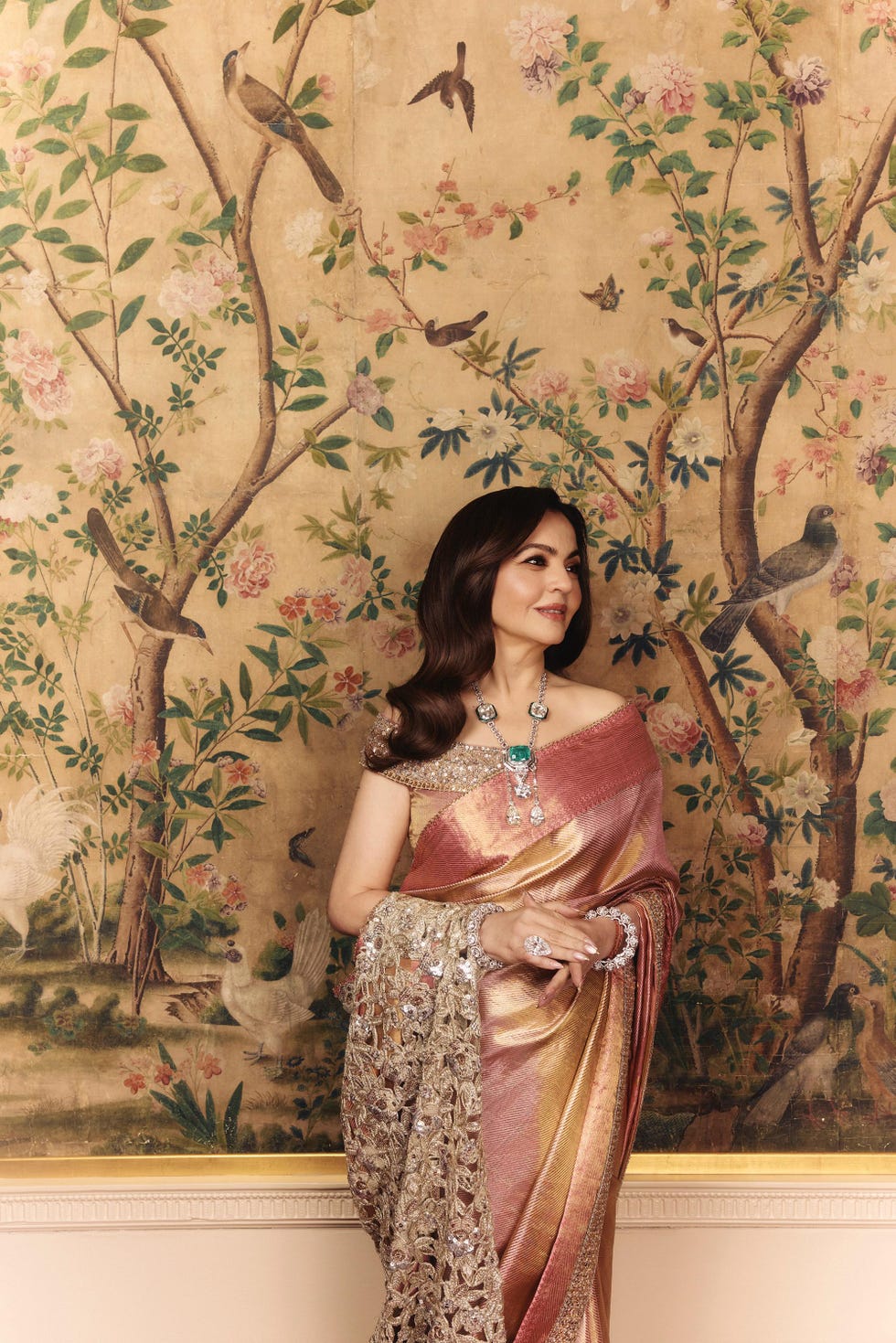 A woman elegantly dressed in a traditional sari stands against a floral wallpaper backdrop featuring birds.