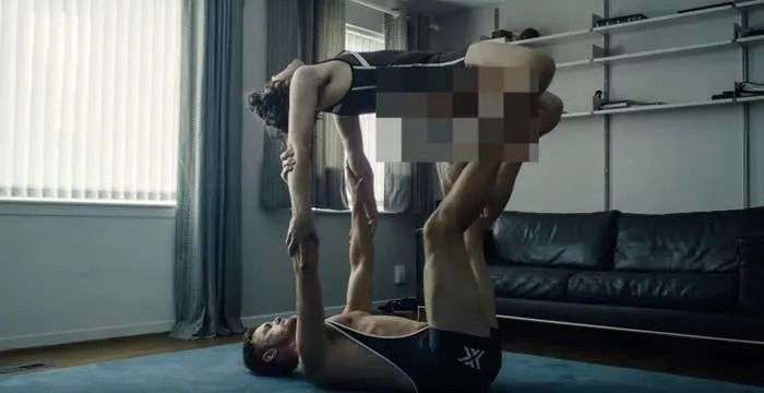 Two people in athletic wear perform an acrobatic pose in a living room, involving balance and coordination