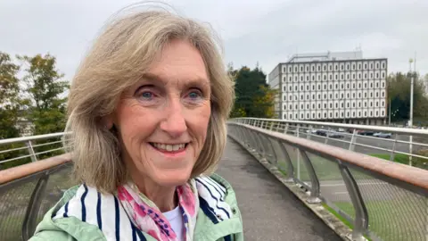 BBC Maureen Millar pictured at a bridge leading to Marlborough House. She has shoulder length light hair and is wearing a green coat with a pink patterned scarf. She is smiling. 