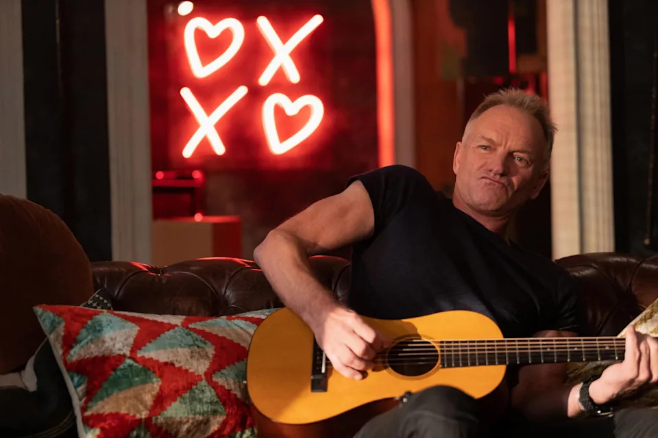 Sting sits on a couch playing a guitar with a neon sign of hearts and XO in the background