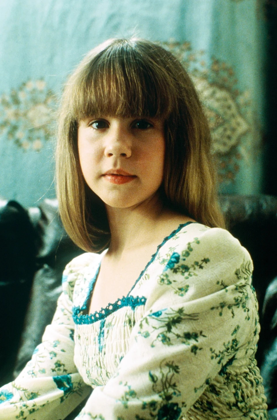 Young girl with long hair, wearing a floral dress, sits on a couch with a calm expression. Background features a detailed pattern