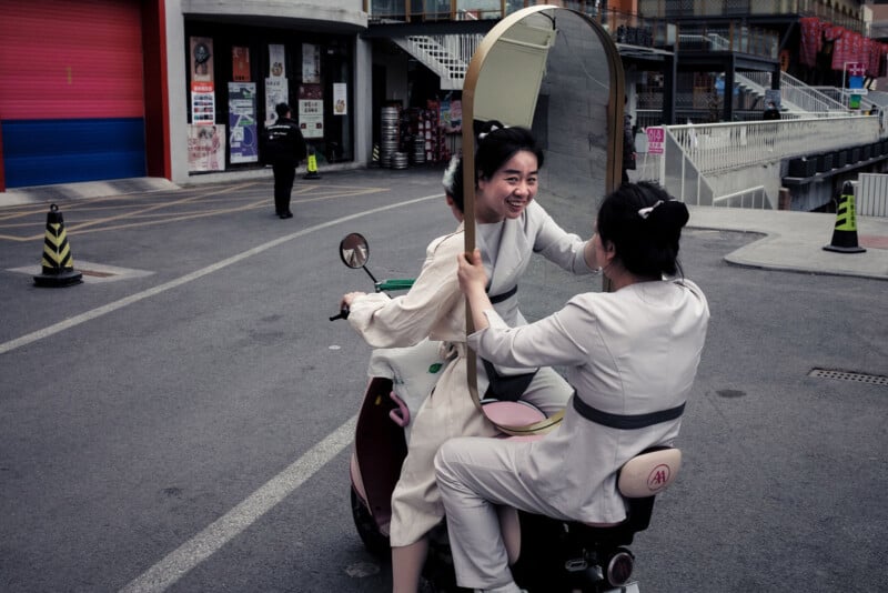 Two people wearing light-colored outfits ride a scooter on a city street. The passenger holds a large mirror facing the driver, reflecting their smiling face. Urban buildings and street signs are visible in the background.