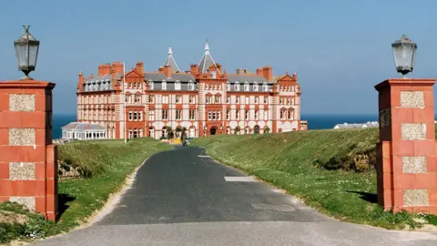 Historic England archive A large, rectangular red and white brick building with several chimneys and pointed triangular roofs stands at the bottom of a grey surfaced driveway with grassed areas to either side. A blue sky and the sea are in the background. In the foreground is the driveway entrance with two red brick pillars each topped by an old-fashioned glass lantern.
