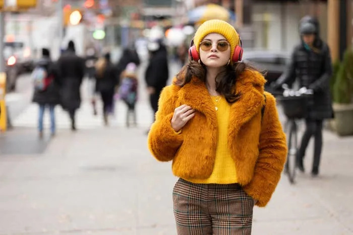 Young woman in a fur jacket, plaid pants, yellow knit hat, sunglasses, and red headphones walking on a city sidewalk
