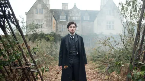 AJ Pics/Alamy Daniel Radcliffe stares into the distance, dressed in a black suit, waistcoat with a chain across the middle and overcoat. He is in an foggy, overgrown garden in front of an old grey stone building. To his sides are the edges of what looks like wrought iron gates that have fallen off their hinges.
