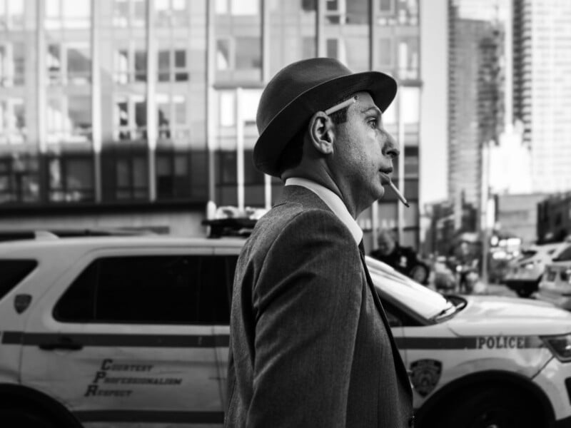 A man in a suit and hat walks past a police car on a city street, with a cigarette in his mouth. Tall buildings are reflected in the windows behind him. The photo is in black and white.