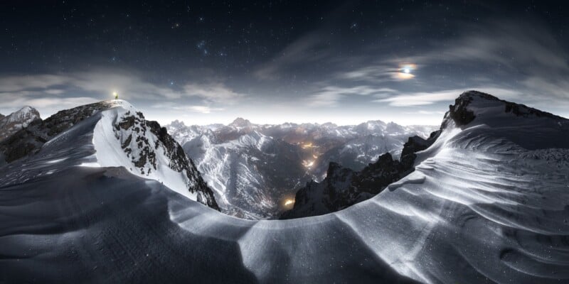 Snow-covered mountain peaks under a starry night sky, with distant towns glowing in the valley below and a bright light atop one peak. Wispy clouds and moonlight illuminate the scene.