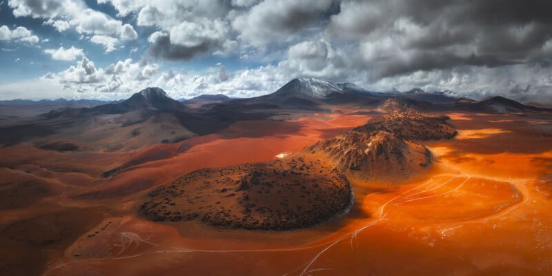 A dramatic landscape of a vast, red desert with scattered dark rocky hills, tire tracks, and distant mountains under a dynamic, cloudy sky with patches of sunlight illuminating the terrain.