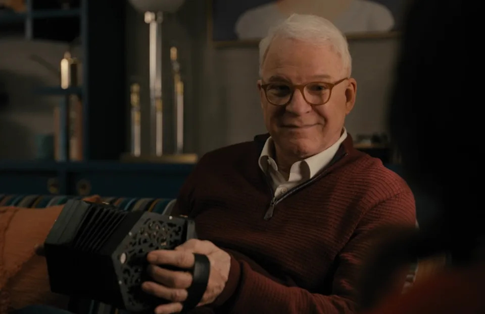 Steve Martin smiles while holding a concertina in a cozy indoor setting