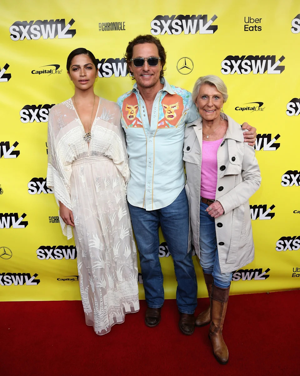 Three people posing on a red carpet at SXSW. The center person is in a casual shirt with western embroidery; the others are in a dress and jacket