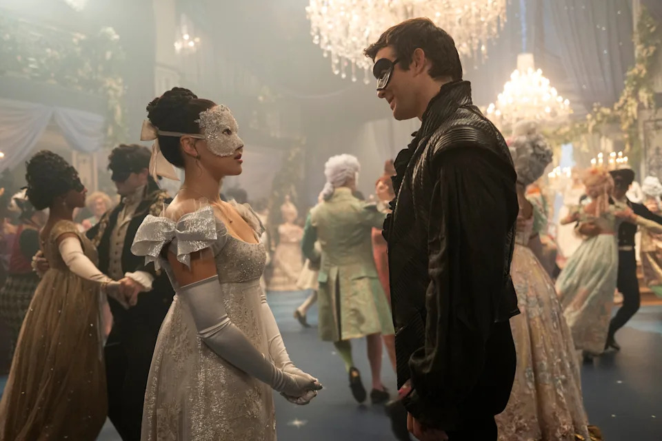 Masked couple in elegant attire faces each other at a grand ballroom, surrounded by dancing guests wearing historical costumes