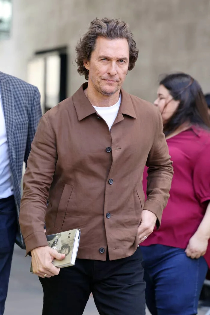 Man in a brown jacket holding a book, walking alongside others in casual settings