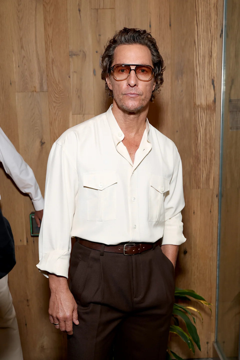 Man in casual-style shirt and pants, wearing large sunglasses, stands against a wooden backdrop