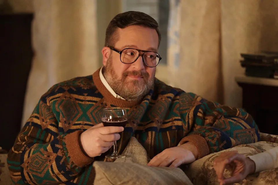 Nathan Lane sits on a sofa, wearing glasses and a patterned sweater, holding a wine glass