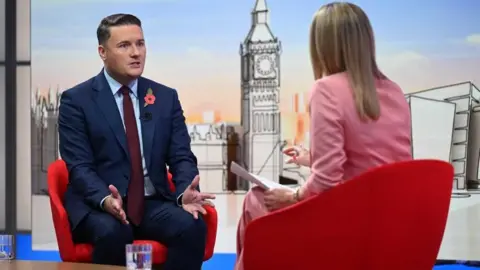 BBC Wes Streeting, whose short brown hair is combed to the side, gestures with his hands and has a serious expression as he wears a navy suit with blue shirt and dark red tie in the Laura Kuenssberg studio