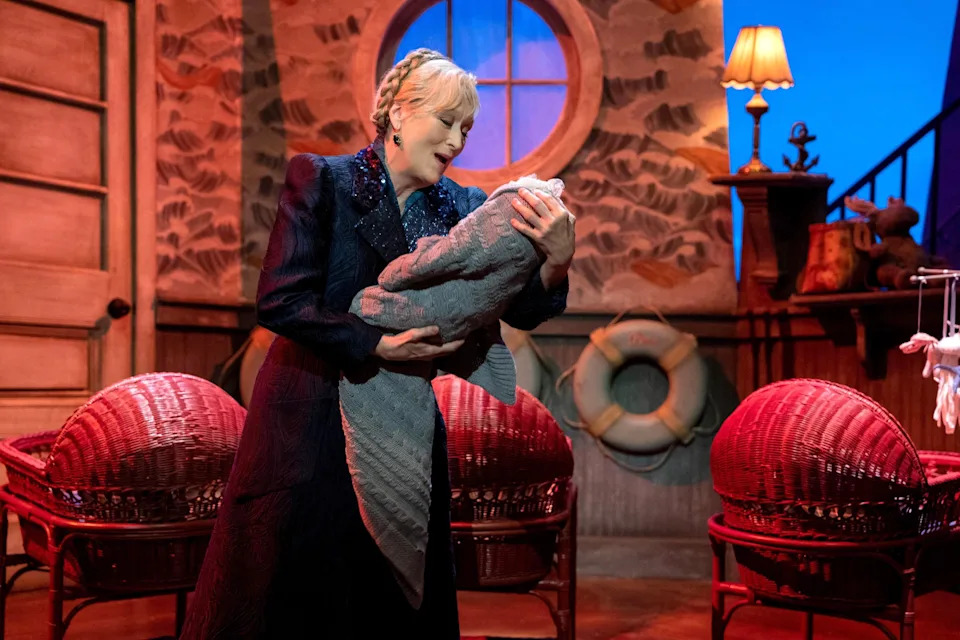 Meryl Streep holds a swaddled doll in a theatrical setting with wicker cribs and nautical decor, suggesting a dramatic performance