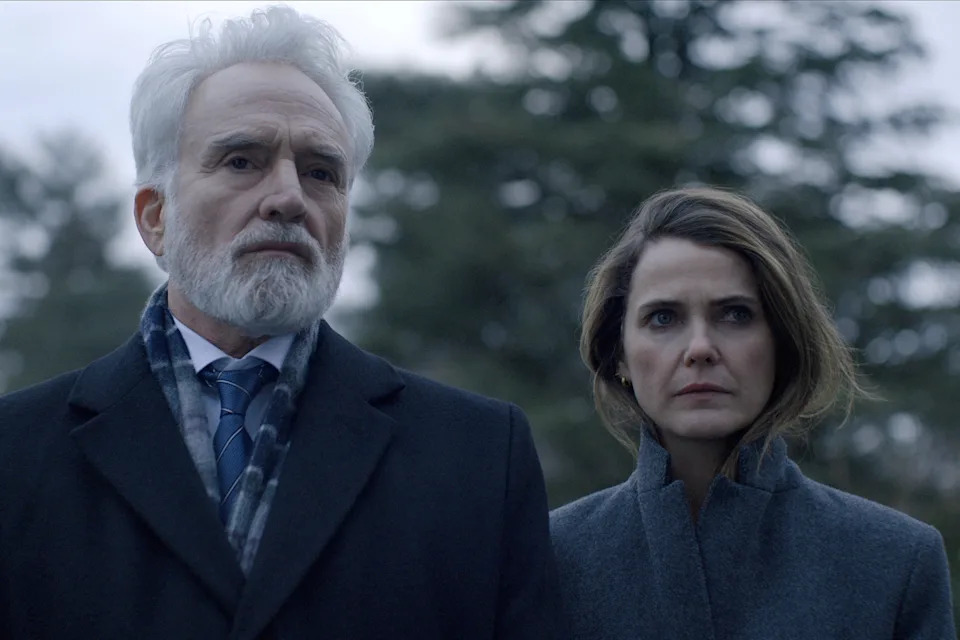 Man with white beard and suit stands beside woman with long hair in dark coat, both outdoors with serious expressions. Trees in the background