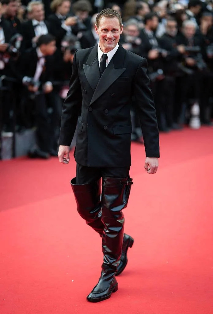 Man on red carpet wearing a double-breasted suit and unique high-shine boots, smiling for the cameras