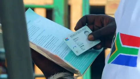 A close-up of a hand holding a South African ID card and checking it against details in a booklet. The sleeve of a T-shirt with the South African flag can also be seen.