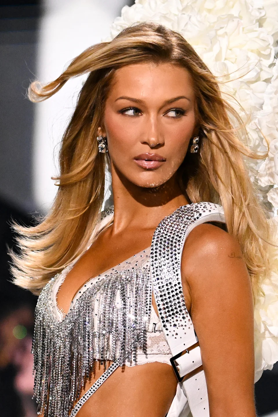 Bella Hadid on runway wearing sparkly, fringed outfit with structured shoulder details, hair flowing, looking sideways