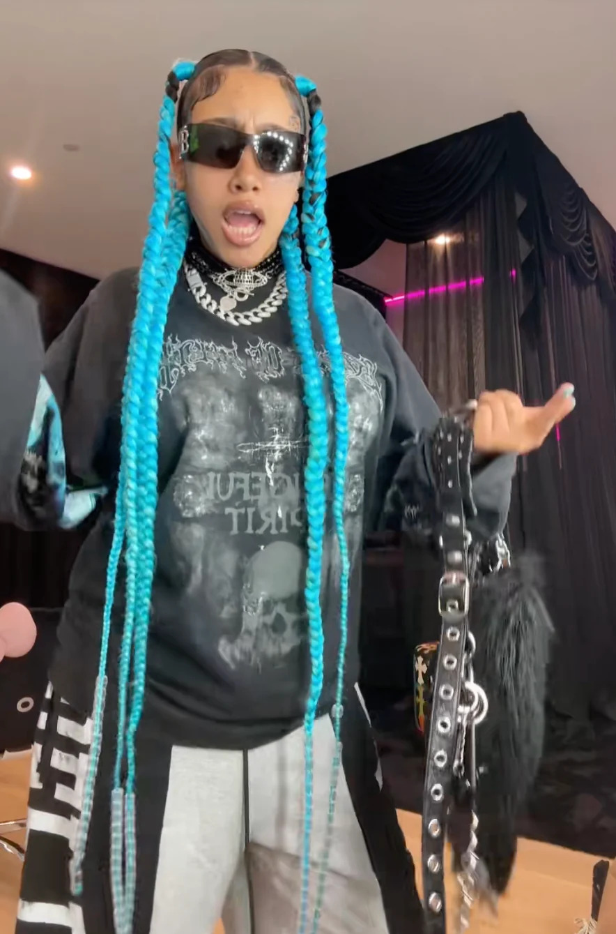 North West wearing sunglasses, a graphic shirt, and accessorized with braided hair poses energetically in a stylish setting