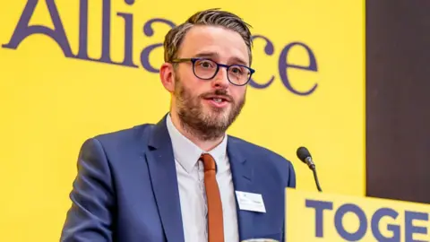 Alliance Party Nick Matheson has a grey beard, grey/whitish hair that is gelled into a spike and grey round glasses. He is wearing a navy shirt, white shirt and orange tie, and behind him is the yellow white Alliance banner