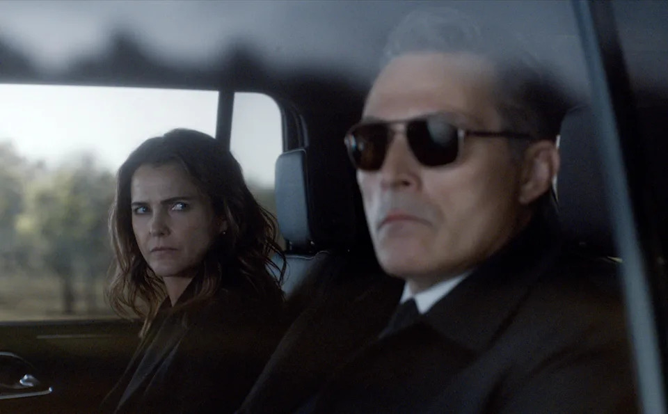 Two people in a car, one wearing sunglasses and a suit, the other with a serious expression