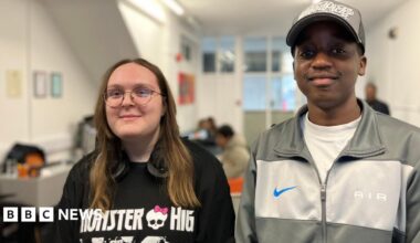 A young woman and a man wearing casual stand next to each other, there are people sitting at table sin the background