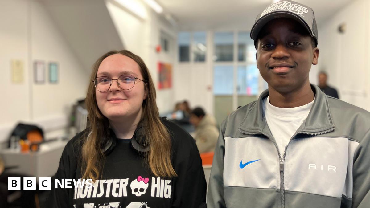 A young woman and a man wearing casual stand next to each other, there are people sitting at table sin the background