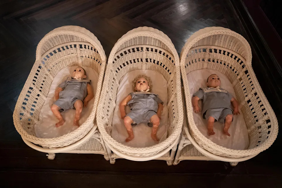 Three baby dolls lie in three wicker bassinets, each dressed in gray outfits, resembling props or display items from a movie or TV show set