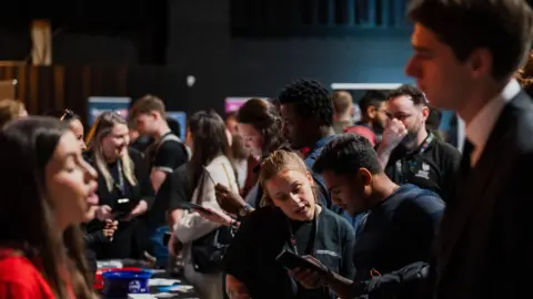 Darogan A man is talking to a woman over a table at a jobs fair while in the background people talk to each other