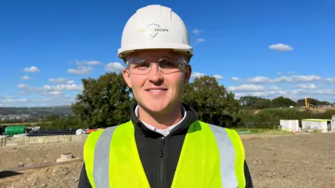 Jack Little on a construction site, wearing a high vis jacket, hard hat, and protective glasses. 