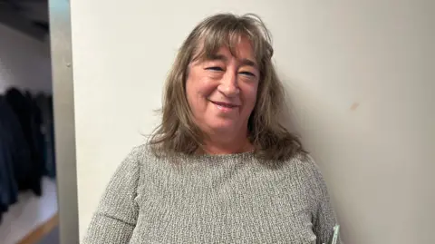 A woman with long, blonde hair, wearing a grey jumper, standing against a cream wall