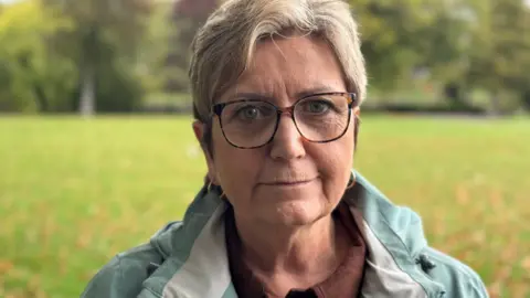 BBC A woman with short blonde hair and wearing tortoiseshell-rimmed glasses looks into the camera without smiling. The collar of a light turquoise coat can be seen around her shoulders and she has two gold ring earrings.