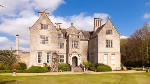 Haakon Dewing A grand, faded brown brick building stands in front of a mowed green lawn. There is an arched doorway and steps leading up to it, trimmed hedges and a tree to the right. The sky is blue with white clouds, the weather sunny.