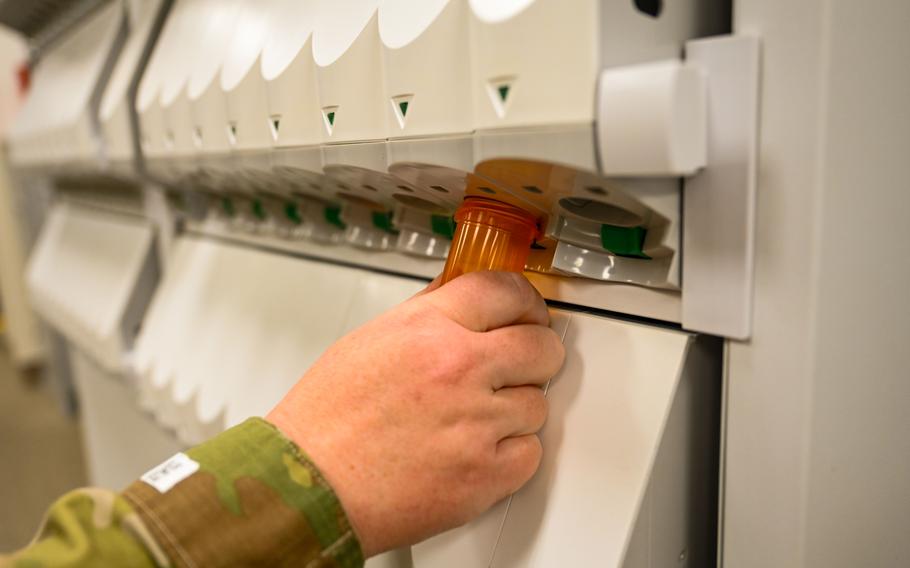A close-up view of a pharmacist’s hand holding a small orange pill bottle up to a dispensing machine.