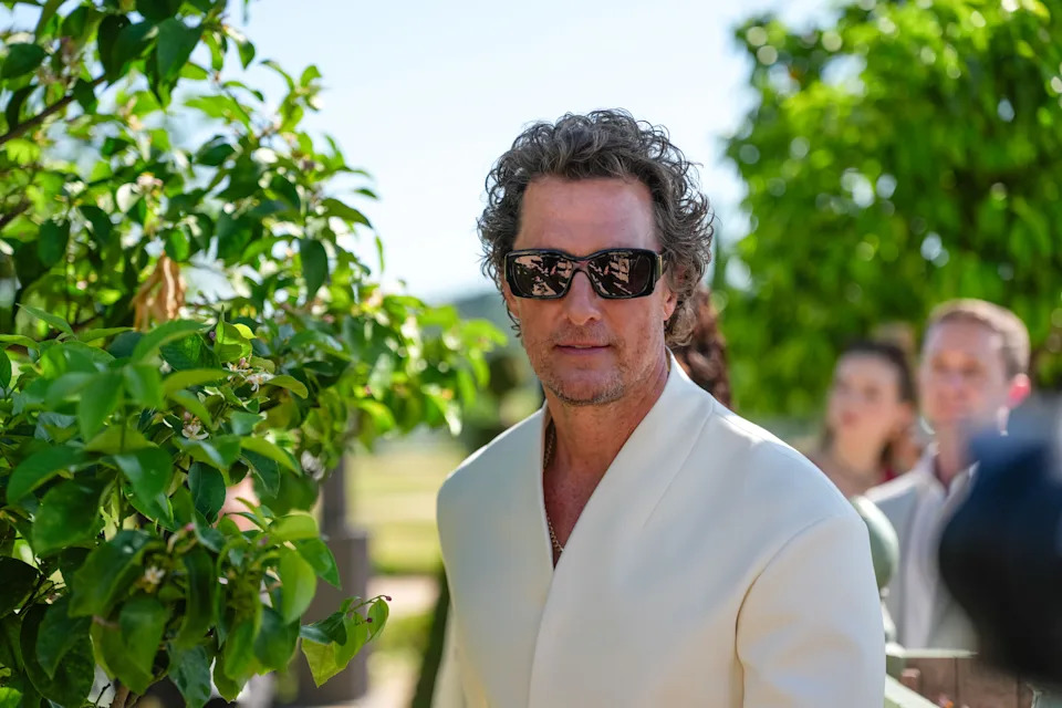 A person with wavy hair and sunglasses wears a stylish white blazer outdoors, surrounded by greenery and people in the background