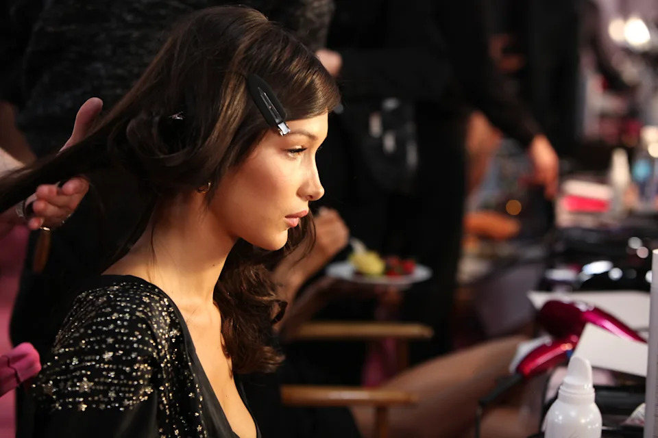 Bella Hadid backstage preps with hair clips and sequined outfit, seated among hair tools and staff in a bustling fashion show environment