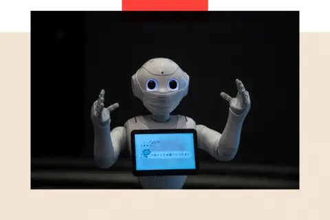 AFP via Getty Images A greeting robot with its two arms in front, with a mask on and a screen on its front 


